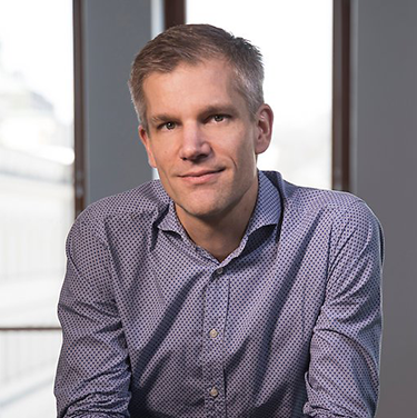 Mattias Blomberg in a purple shirt leaning towards the camera in front of a grey wall and some windows